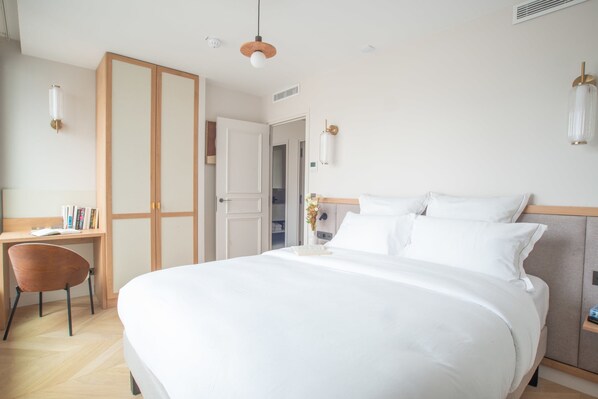 A hotel room with a large bed, a desk with a chair, a wardrobe, and a bedside table with a lamp.