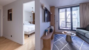 A modern hotel room with a bed, a seating area with books, a TV, and a balcony with a view.