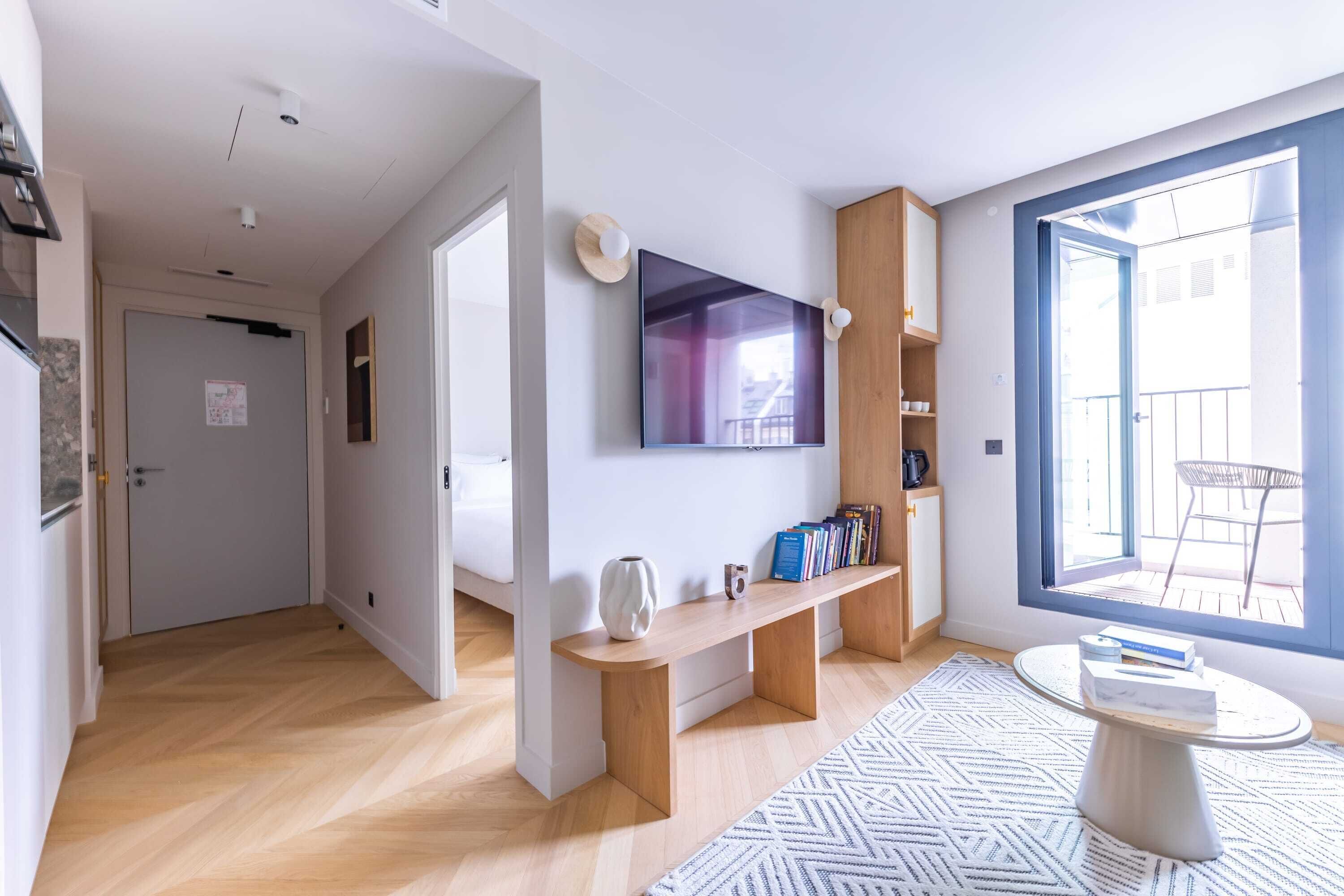Una sala de estar moderna con un televisor de pantalla plana, estanterías de madera, una mesa de centro redonda y un ventanal grande.