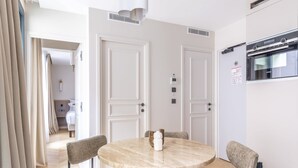 A modern hotel room with a round dining table, four chairs, a built-in oven, and a separate bedroom visible through an open door.