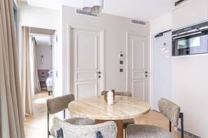A modern hotel room with a round dining table, four chairs, a built-in oven, and a separate bedroom visible through an open door.