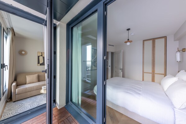 Une chambre moderne dotée d’un grand lit, d’une porte-fenêtre coulissante donnant sur un balcon et d’un espace de vie séparé avec un canapé.