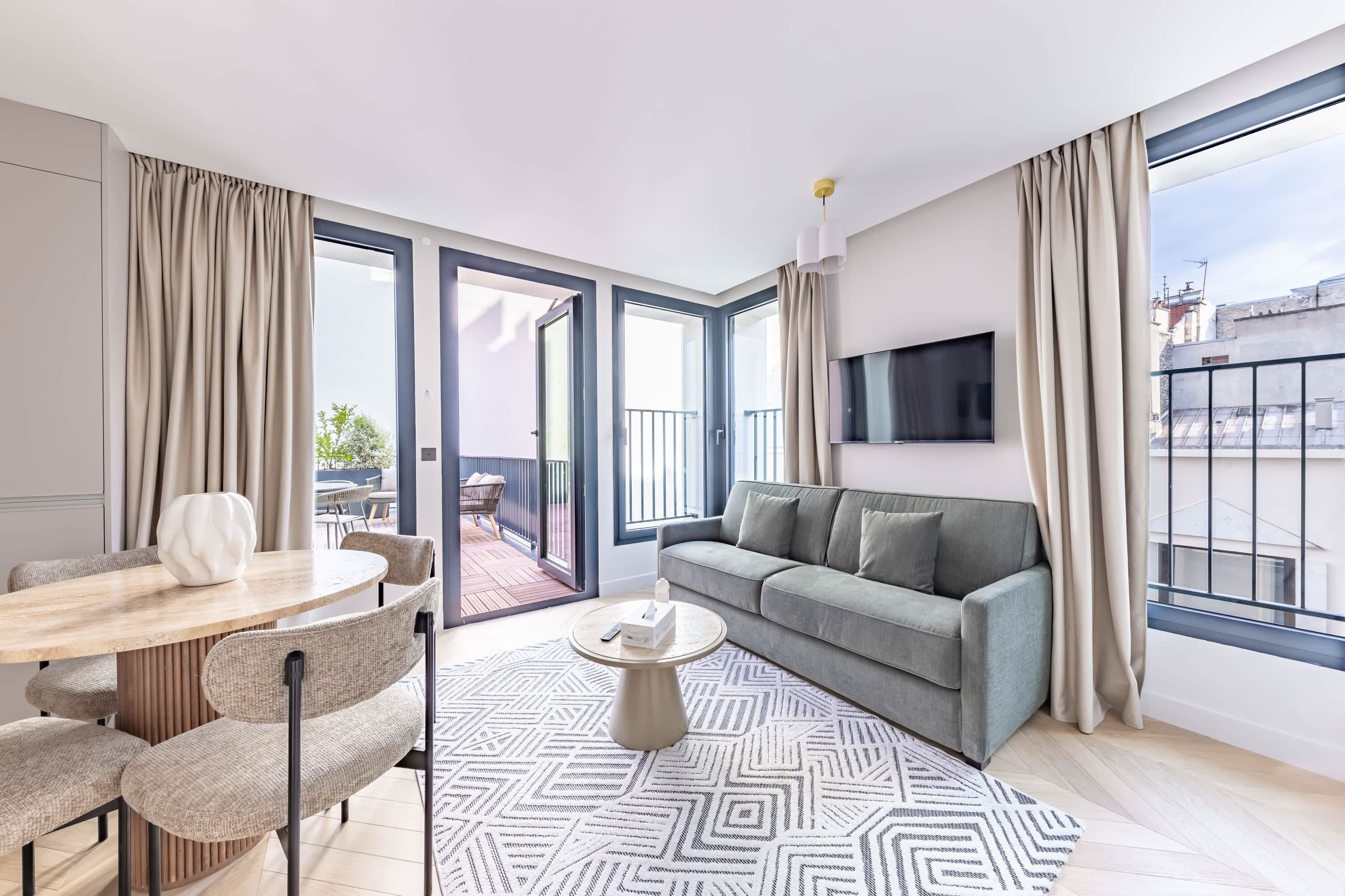 A modern living room with a sofa, a round coffee table, a dining table with chairs, and a large window with curtains.