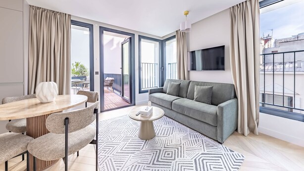 Een moderne woonkamer met een bank, een rond bijzettafeltje, een eettafel met stoelen en een groot raam met gordijnen.