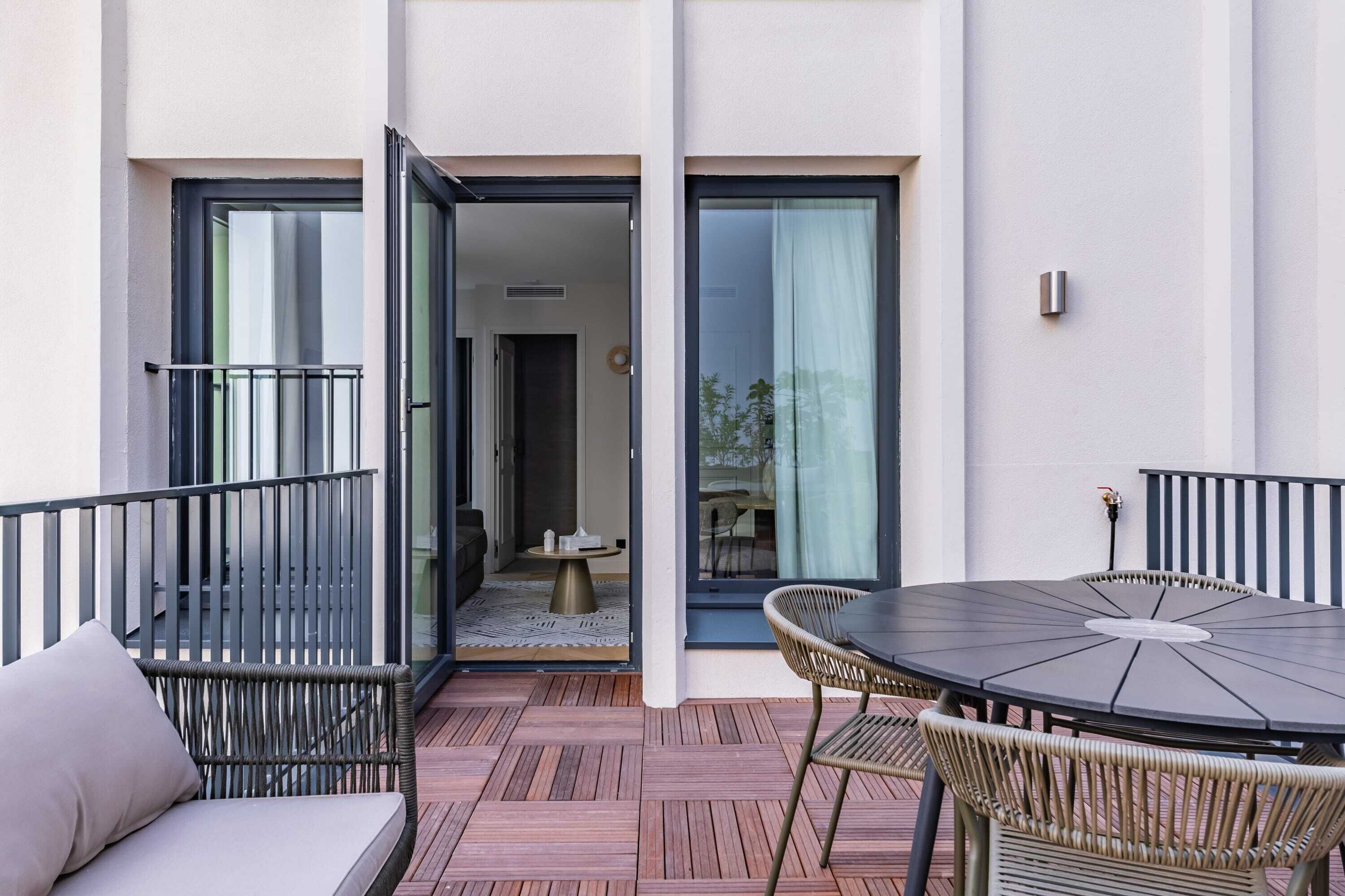 Un patio moderno al aire libre con una mesa redonda y sillas de mimbre, una pequeña mesa redonda con una vela, y un ventanal corredero que da a un espacio interior.