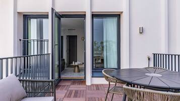 Un patio moderno al aire libre con una mesa redonda y sillas de mimbre, una pequeña mesa redonda con una vela, y un ventanal corredero que da a un espacio interior.