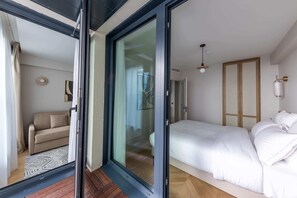 A modern bedroom with a bed, a glass door leading to a balcony, and a sofa in the adjacent room.