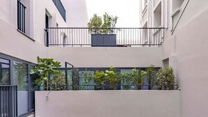 Une petite cour intérieure en milieu urbain, dotée d’une rambarde en verre, de plantes en pot et d’un portail en métal.