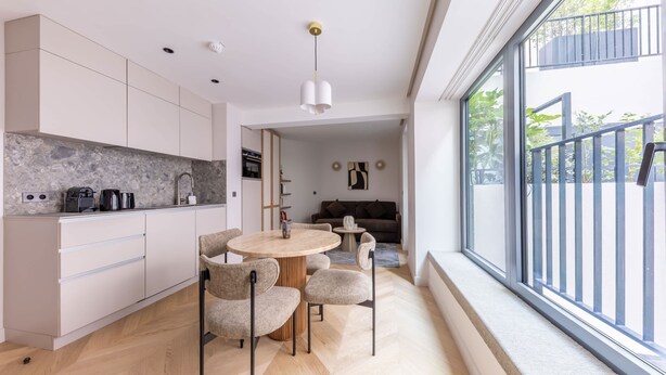 Une cuisine moderne avec des meubles blancs, un coin repas doté d’une table ronde et de chaises, et une grande fenêtre donnant sur un balcon.