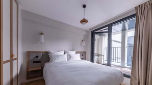 A modern hotel room with a large bed, a built-in nightstand, a wall-mounted lamp, and a sliding glass door to the balcony.