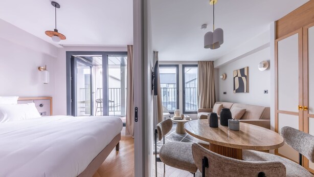A modern hotel room with a large bed, a round dining table with chairs, a sofa, and a view of the city through large windows.