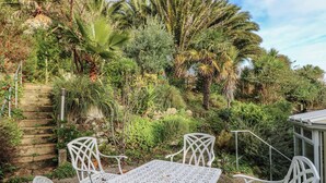 A garden patio with white metal chairs and a table, surrounded by lush greenery and palm trees. - Tall Timbers (Ventnor)