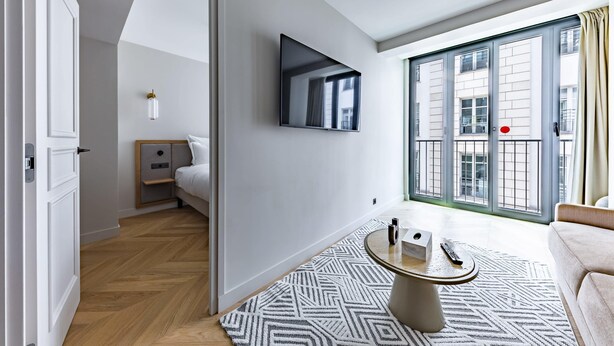 A modern hotel room with a bed, a TV mounted on the wall, a small bedside table, a round coffee table, and a large window with a view of buildings.