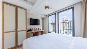 A hotel room with a bed, a desk with a TV, a bookshelf, and a balcony with a city view.