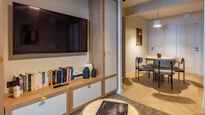 En moderne stue med et fladskærms-tv, et træsofabord med bøger og pyngeting, et rundt sofabord med et magasin, og et spiseområde med et rundt bord og stole.