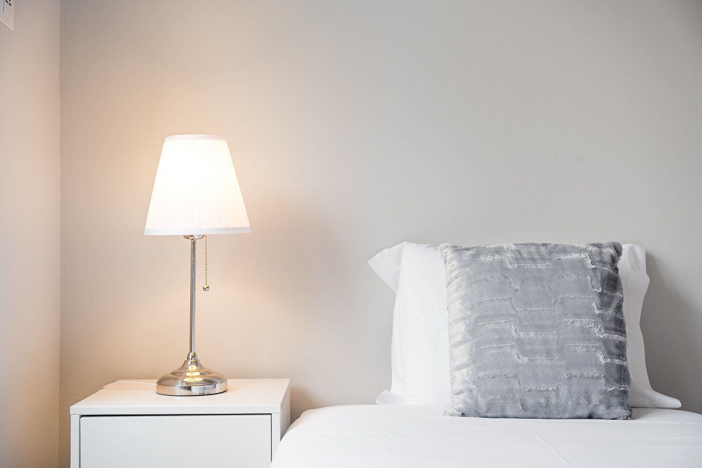Een netjes opgemaakt bed met een wit kussen en een grijs kussen met textuur, een witte nachtkastje met een lamp, en een effen muur.