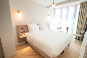 Quarto de hotel moderno com cama grande, mesas de cabeceira, uma cadeira e janela com cortinas.