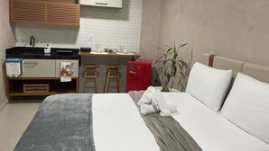 Une chambre d’hôtel compacte comprenant un coin cuisine, un lit avec du linge de lit blanc, un plaid gris et une petite plante en pot.