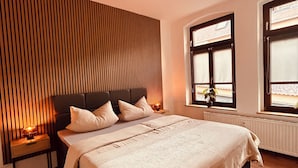 Une chambre avec un grand lit, un mur lambrissé en bois, des tables de chevet avec des lampes et des fenêtres équipées de stores.