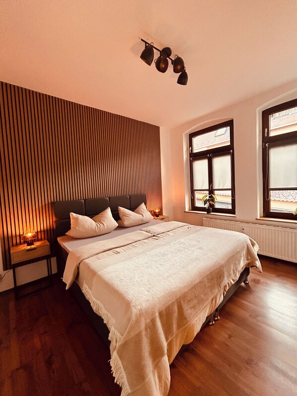 A bedroom with a large bed, wooden panel wall, bedside tables with lamps, and windows with blinds.