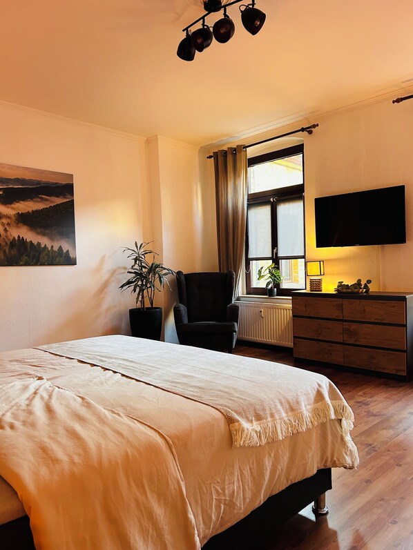 Ein Schlafzimmer mit Bett, Sessel, Fernseher, Fenster mit Vorhängen, einer Pflanze und einem Bild an der Wand.