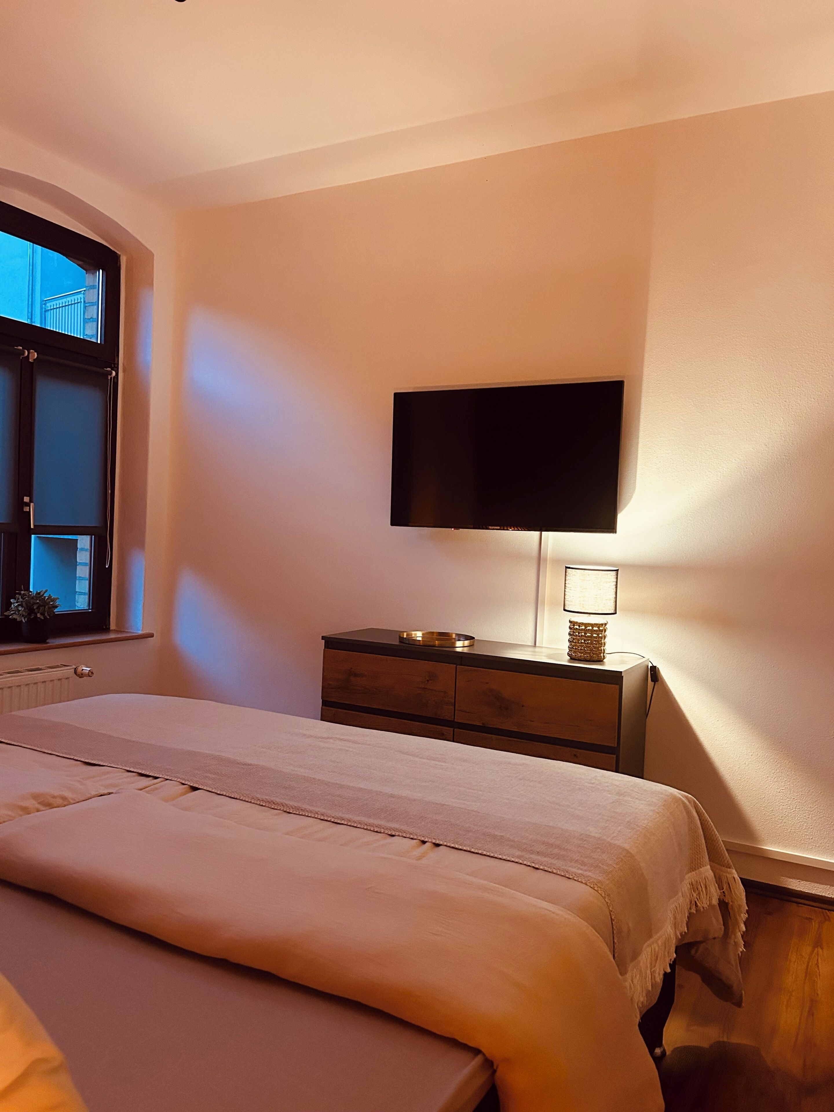 Een slaapkamer met een bed, een aan de muur bevestigde televisie, een bedlamp en een raam met uitzicht op gebouwen.