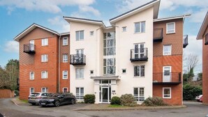 A multi-story residential building with a central entrance, balconies, and a parking area with cars.