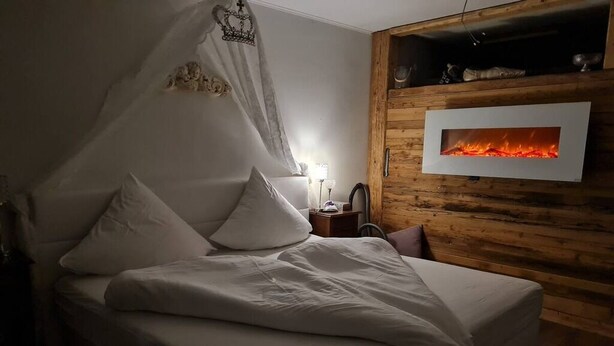 A bedroom with a bed, a bedside table, a lamp, and a wall-mounted fireplace. - Bad Füssing ... time out for two ...  (Bad Füssing)