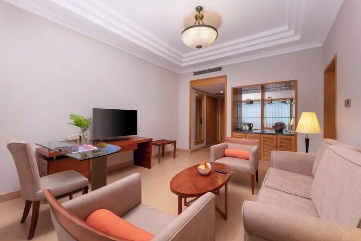 Habitación de hotel con televisión, sofá, sillón, mesa de centro y zona de comedor con mesa de vidrio y sillas.