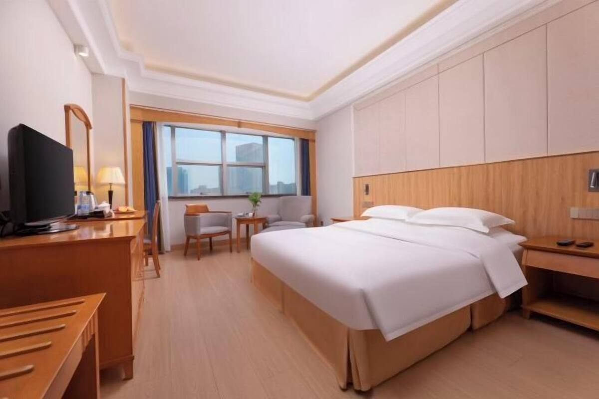 Habitación de hotel con cama, escritorio, silla, televisor y ventana con vistas a la ciudad.
