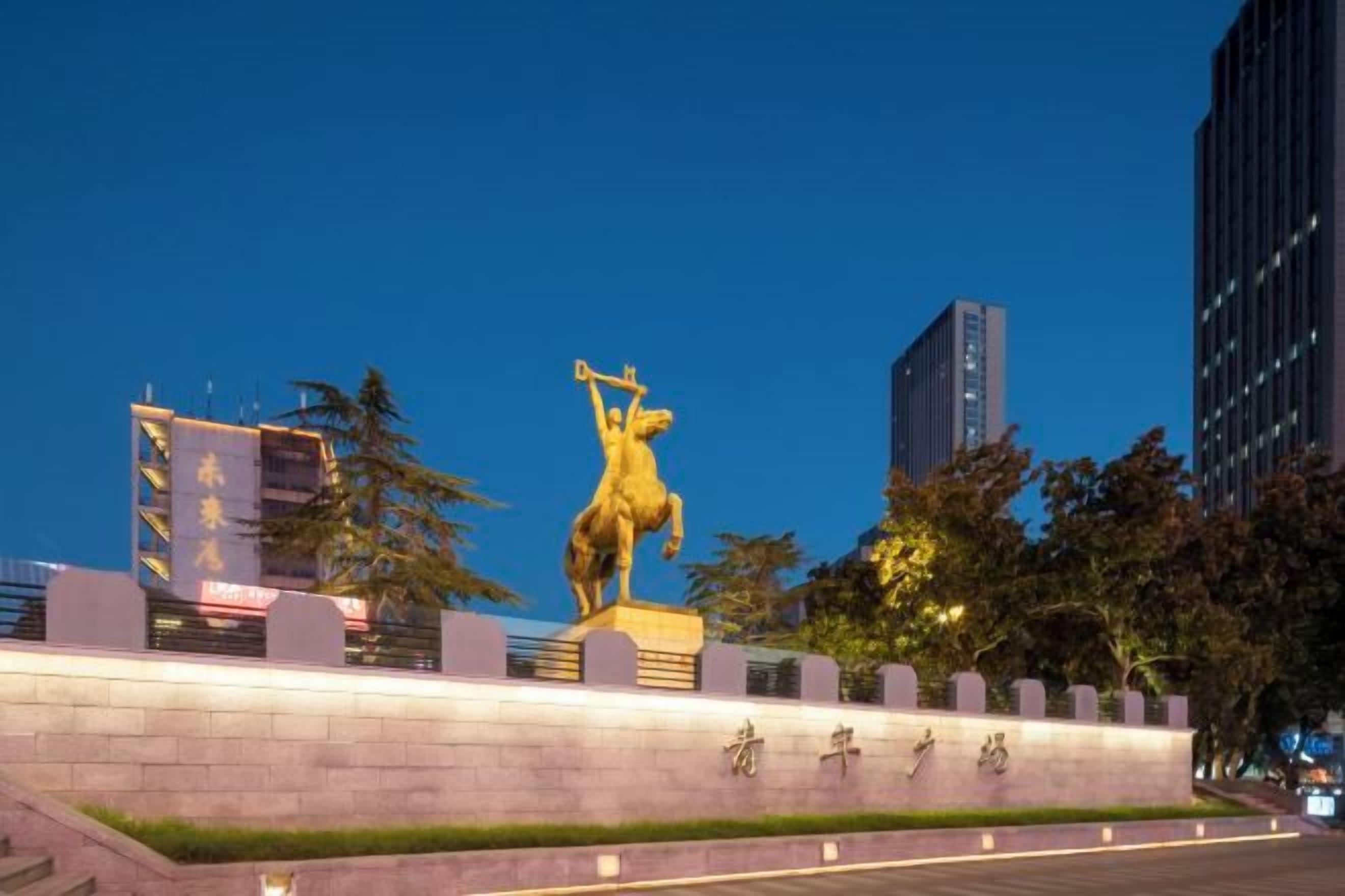 Una estatua de un caballo sobre un pedestal en un entorno urbano, con edificios y árboles al fondo.