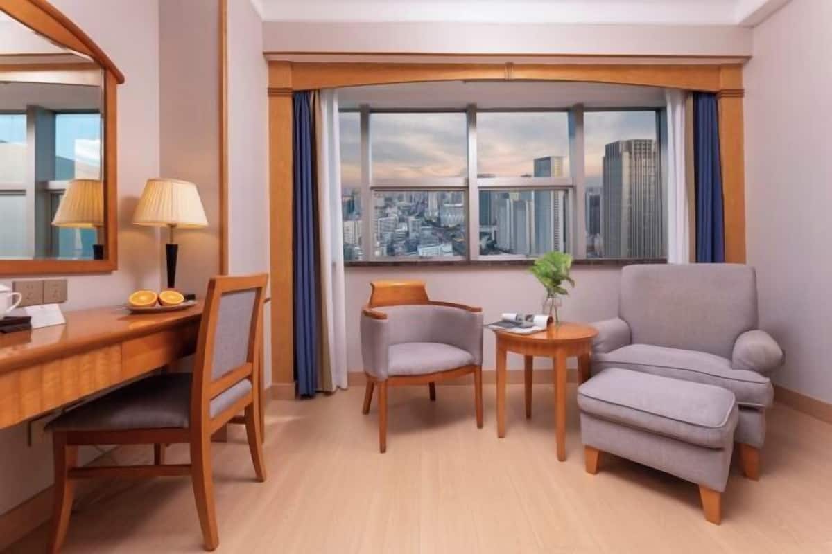 Habitación de hotel con un escritorio de madera, dos sillas, una mesita con un jarrón y un ventanal con vistas a la ciudad.