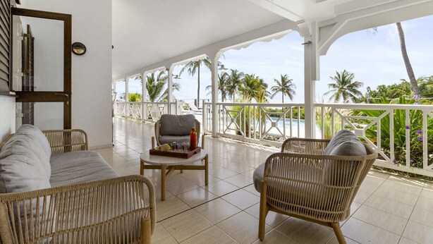 A covered outdoor seating area with wicker furniture, a small table, and a view of palm trees and a pool. - Villa 3 Anses - Three Bedroom Villa, Sleeps 8 (Sainte-Anne)