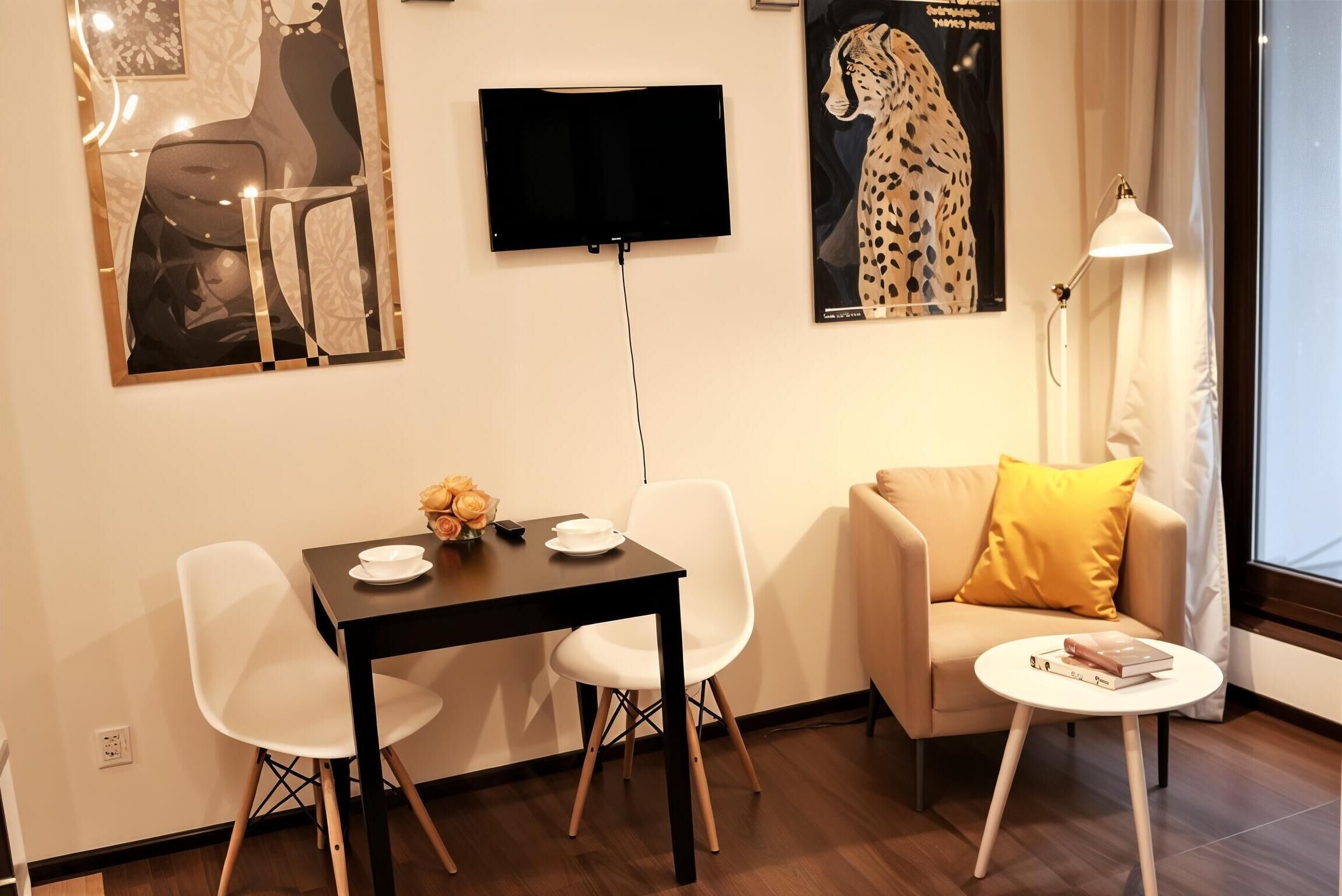 A modern living room with a black TV mounted on the wall, a white sofa with a yellow pillow, a small round table with books, and a dining area with a black table and white chairs.