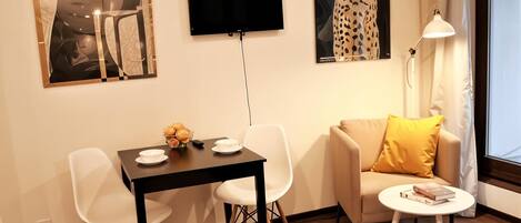 A modern living room with a black TV mounted on the wall, a white sofa with a yellow pillow, a small round table with books, and a dining area with a black table and white chairs.