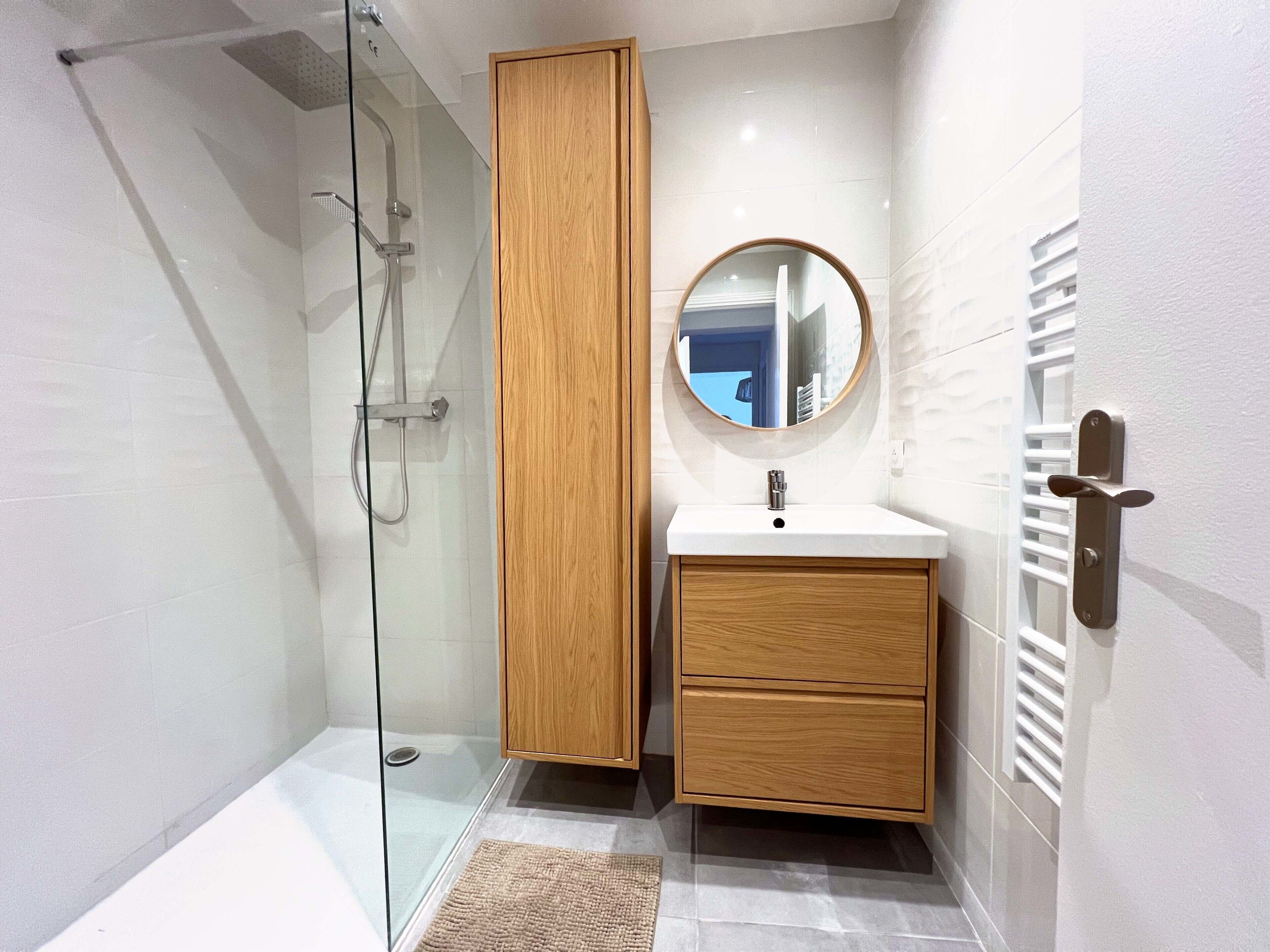 Een moderne badkamer met een douchecabine van glas, een houten kastje, een ronde spiegel en een witte wastafel.