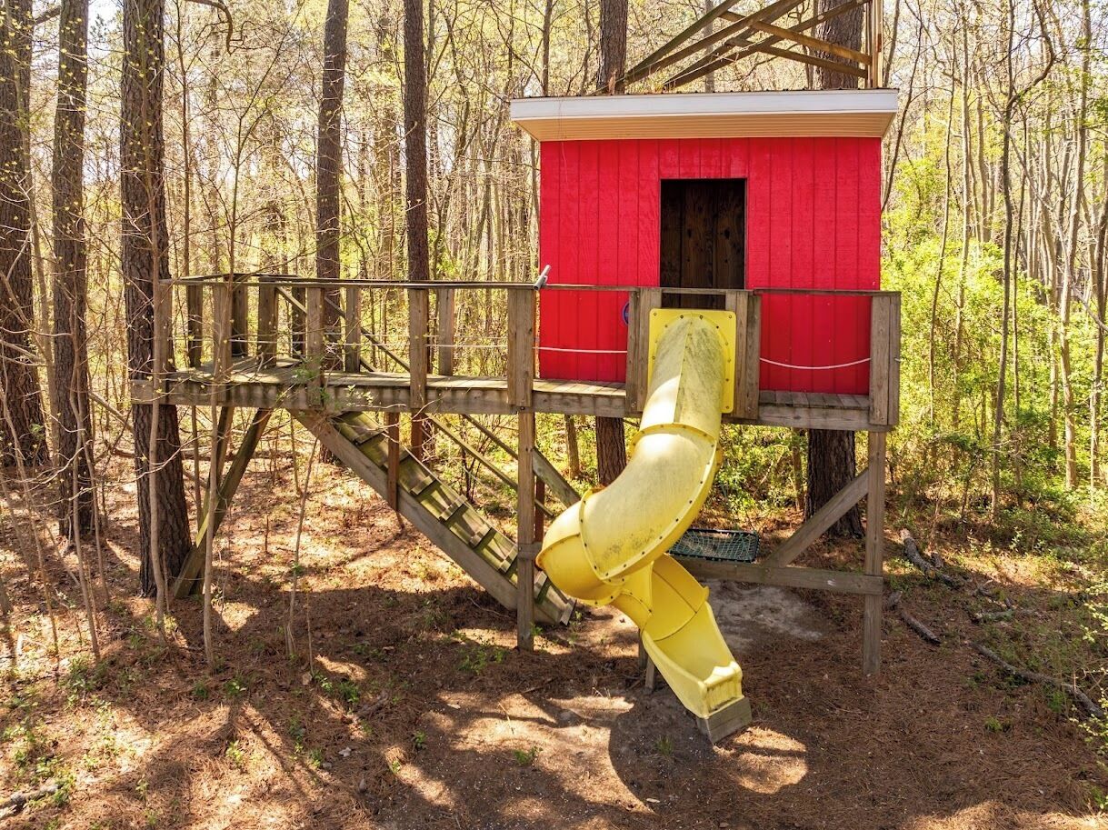 A red wooden playhouse with a yellow slide in a forest setting.
