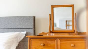 Ein hölzerner Schminktisch mit Spiegel und zwei Schubladen, neben einem Bett mit grauem Kopfteil und weißer Bettwäsche.