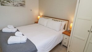 Ein ordentlich bezogenes Bett mit weißen Leinentüchern und einer grauen Tagesdecke, zwei Nachttischlampen, ein kleiner Nachttisch und ein weißer Kleiderschrank.