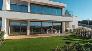 A modern two-story house with large sliding glass doors, a well-maintained lawn, and a patio with outdoor furniture.