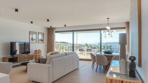 A modern living room with a flat-screen TV, a sofa, a dining area with chairs, and a large window offering a view of the outdoors.