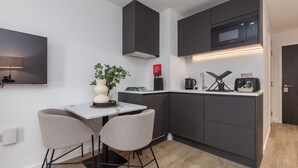 A modern kitchen with dark cabinets, a white countertop, and a built-in microwave. There is a small table with two chairs and a potted plant. - City Living Studio - Studio Apartment, Sleeps 2 (Manchester)