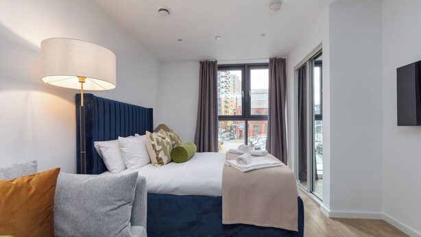 A modern hotel room with a bed, a sofa, a lamp, and a large window offering a city view. - City Living Studio - Studio Apartment, Sleeps 2 (Manchester)