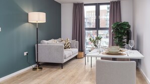 A modern living room with a grey sofa, a floor lamp, a dining table set with plates and glasses, and a large plant.