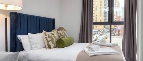 A hotel room with a bed, a lamp, a view of the city through large windows, and a balcony.