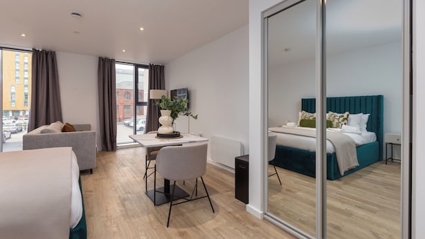 A modern hotel room with a large bed, a desk with a chair, a sofa, and a view of the city through large windows. - Stylish City Studio - Studio Apartment, Sleeps 2 (Manchester)