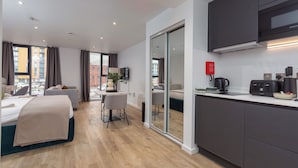 A modern hotel room with a bed, a desk, a chair, a TV, a kitchen area with a microwave, and a sliding glass door leading to a balcony. - Stylish City Studio - Studio Apartment, Sleeps 2 (Manchester)