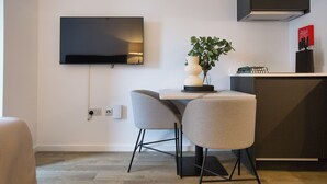 A modern kitchen with a flat-screen TV mounted on the wall, two grey chairs, a small table, and a potted plant. - Elegant Manchester Studio (Manchester)