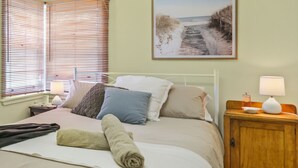 A neatly made bed with pillows, a bedside table with a lamp, and a framed picture of a beach path on the wall. - Coastal Retro House by Book Jervis Bay - Husky (Huskisson)