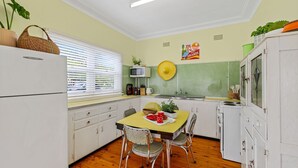 A compact kitchen with white cabinets, a dining table with chairs, a microwave, and a stove. - Coastal Retro House by Book Jervis Bay - Husky (Huskisson)